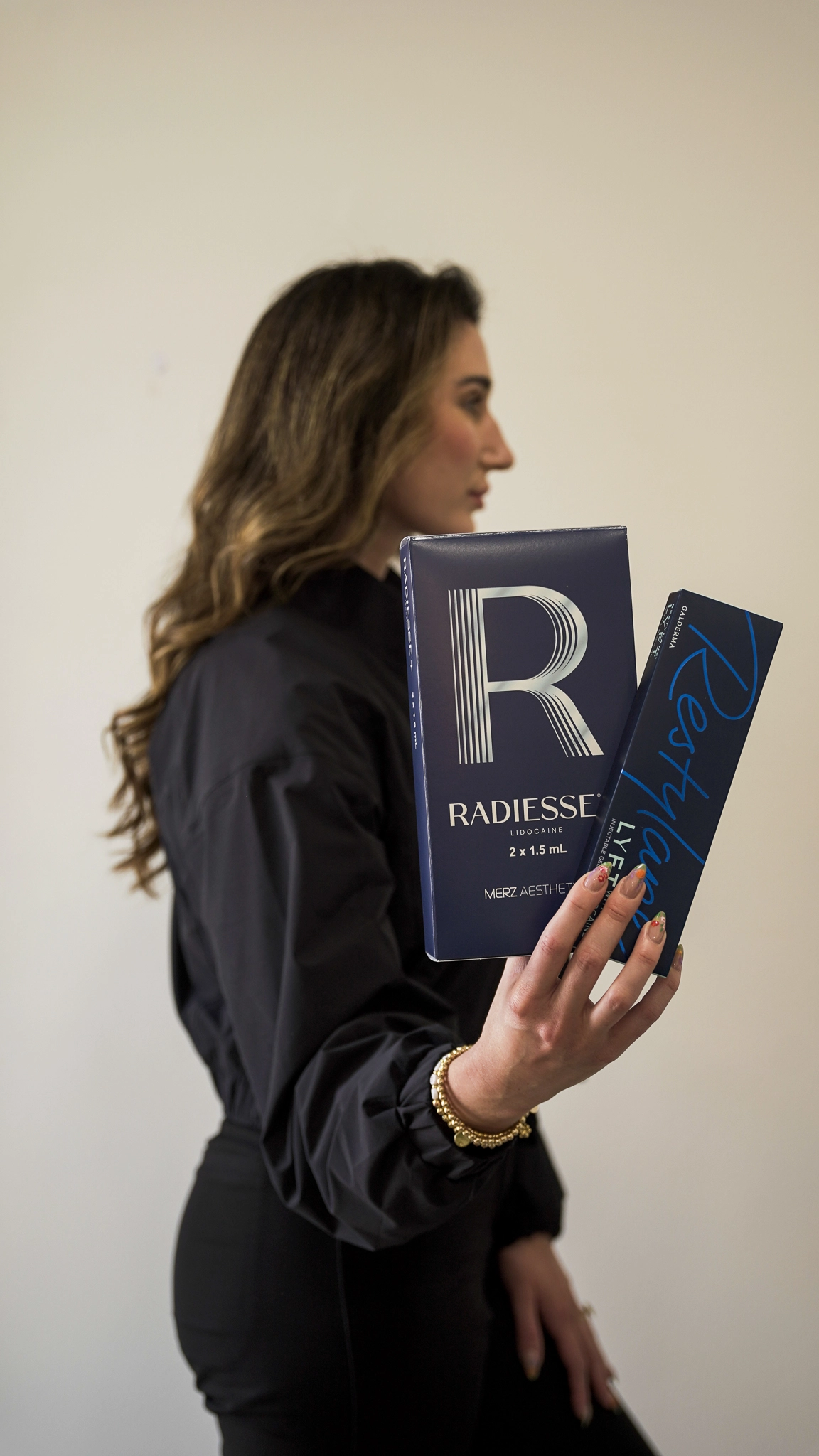 A woman in a black outfit holds Radiesse and Sculptra boxes in profile against a plain light wall.